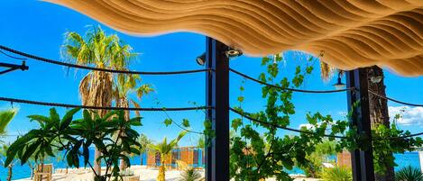 An outdoor seating area with tables and chairs, surrounded by plants and a view of the sea.