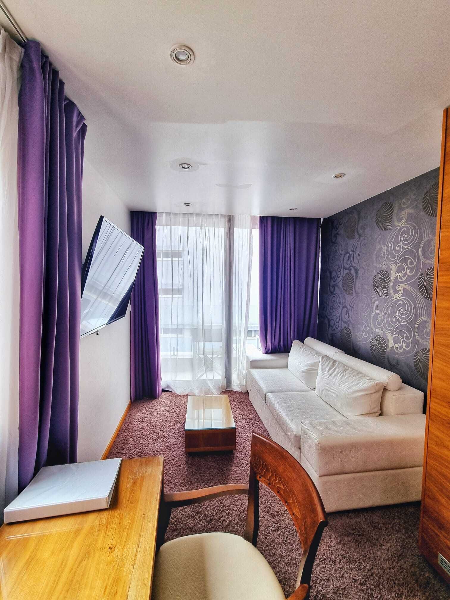 Una habitación de hotel moderna con sofá, una mesa de madera, una silla y una ventana con cortinas moradas.