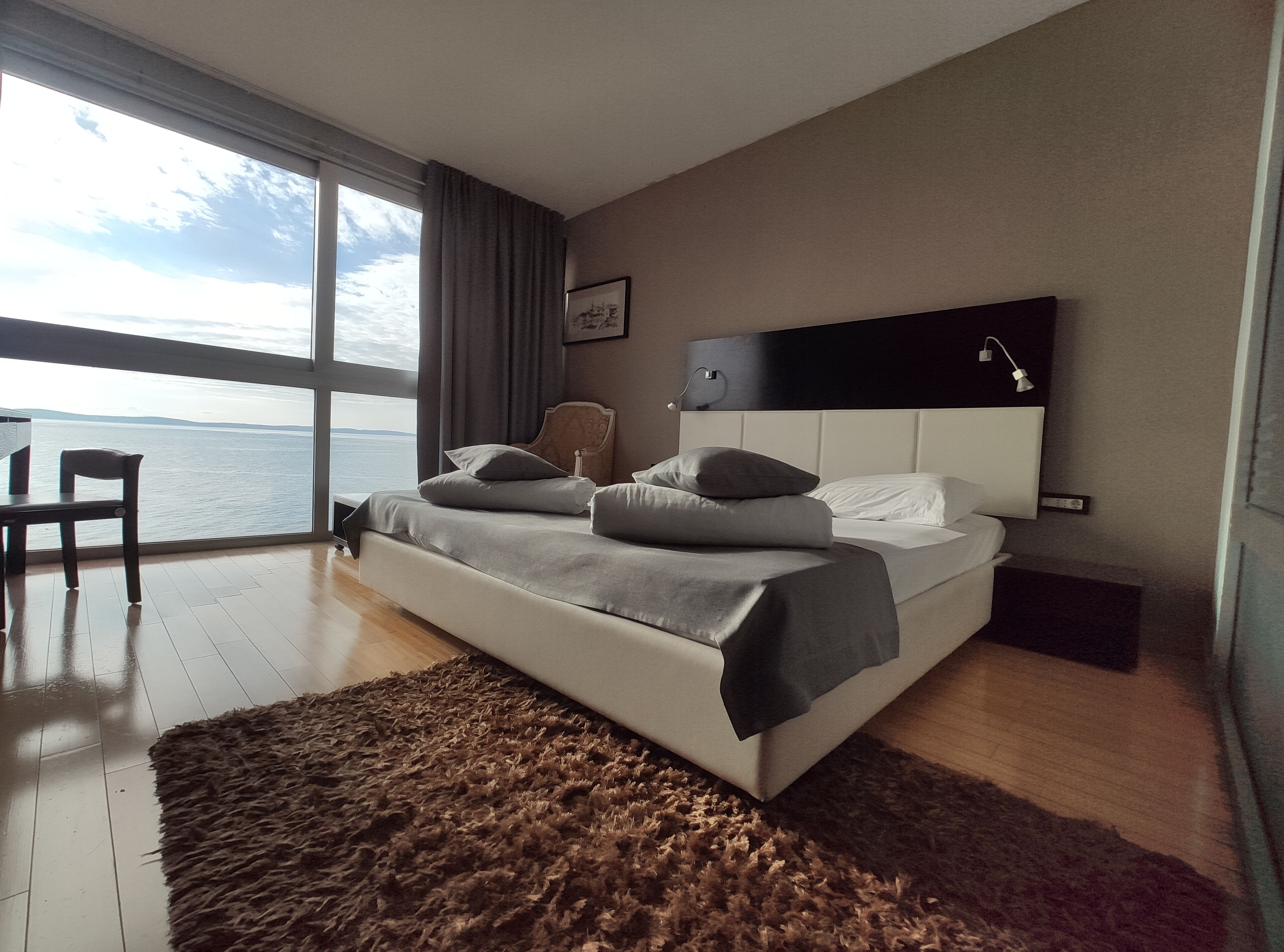 Ein modernes Schlafzimmer mit großem Fenster, einem Bett, einem Sessel und Blick aufs Meer.