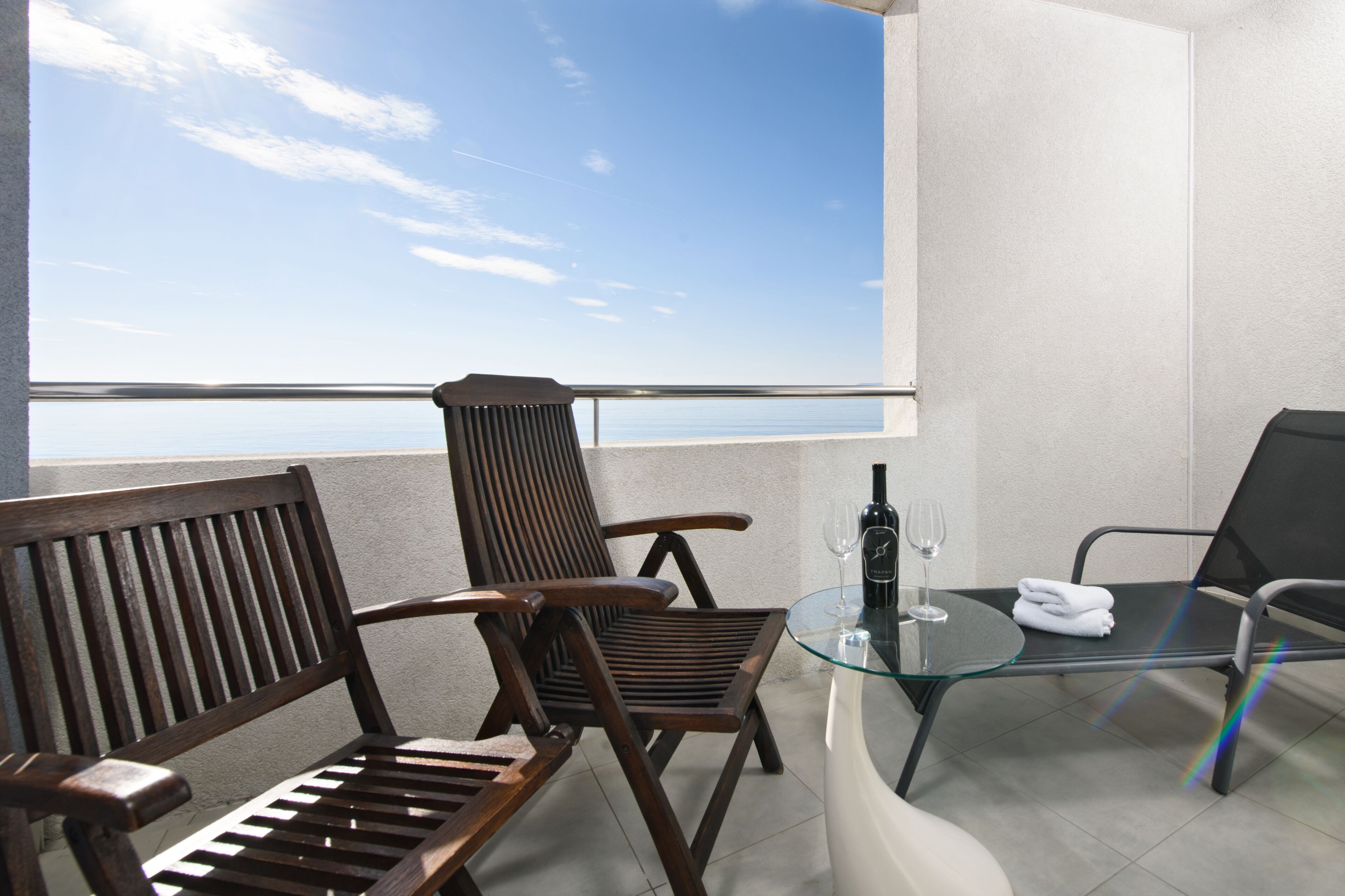 A balcony with a wooden bench, a glass table with a bottle and glass, and a view of the sea.