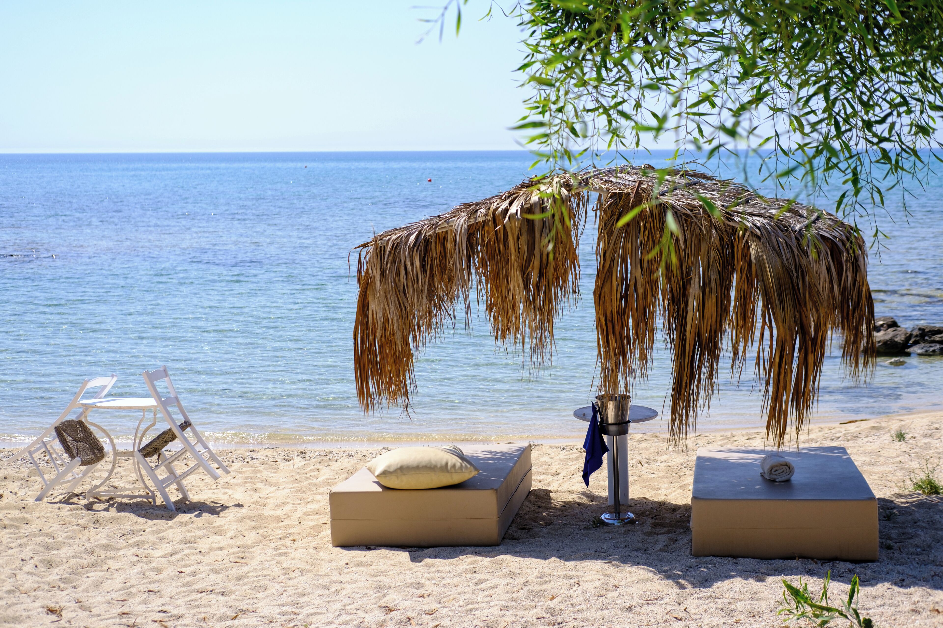 A beach with two lounge chairs, a small table, and a thatched umbrella providing shade.