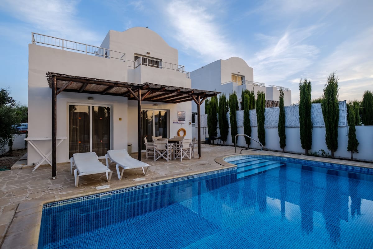 Un área de piscina moderna con una piscina azul cristalina, sillones de descanso y una zona de estar cubierta.