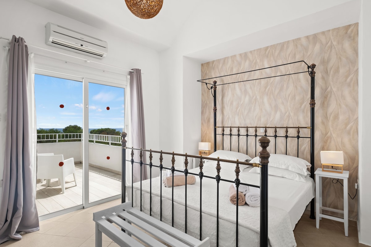 Un dormitorio con cama con dosel, un balcón con sillas blancas y vista al cielo.