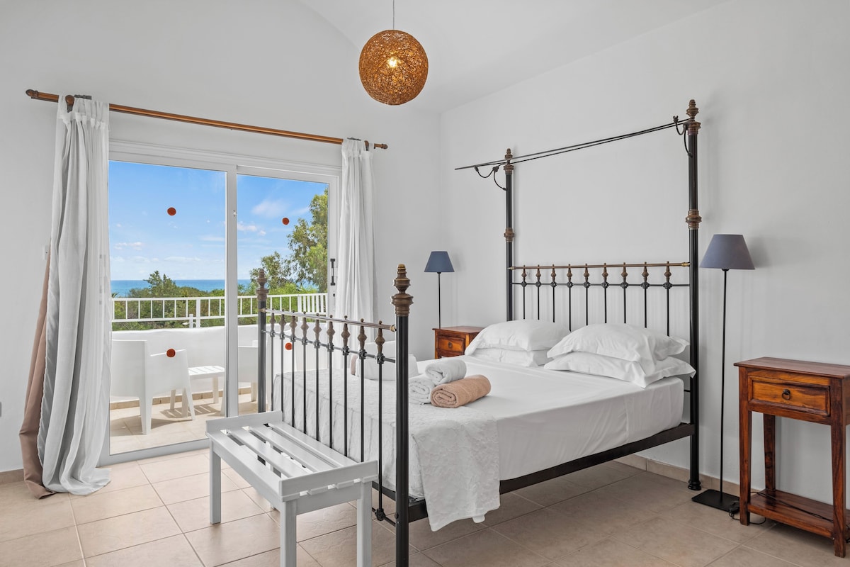 Un dormitorio con cama con dosel, un balcón con una silla blanca y vista al mar.