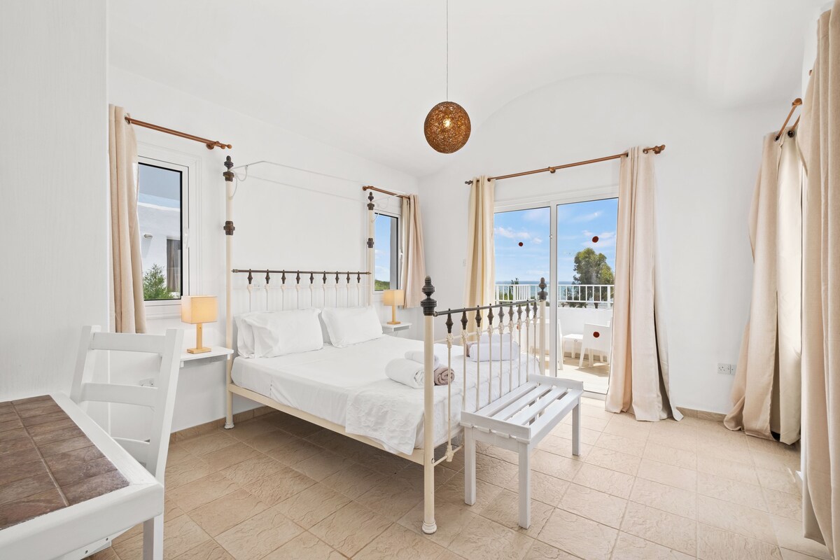 Un dormitorio con cama, mesitas de noche, una silla, un balcón con vistas y cortinas.