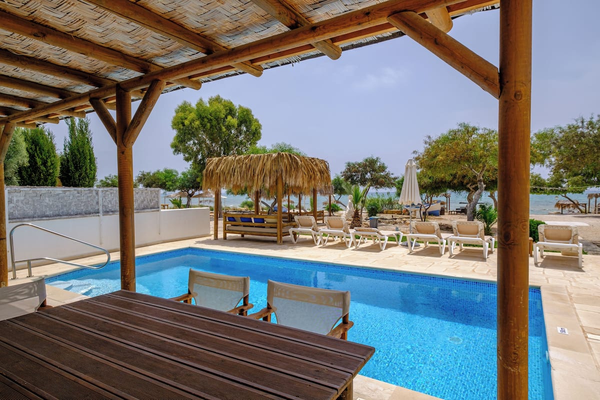 Un área de piscina con una pérgola de madera, sillones de descanso y un parasol de paja.
