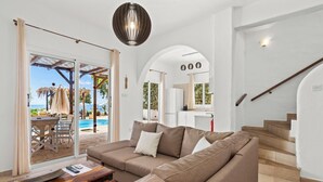 A modern living room with a large beige sectional sofa, a wooden coffee table, and a glass door leading to an outdoor pool area. - Thalassines Beach Villas (Sotira)