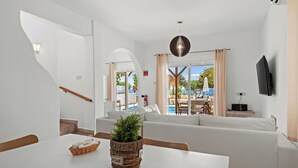 A modern living room with a white sofa, a dining table with a basket and a plant, a TV mounted on the wall, and a view of an outdoor pool area. - Thalassines Beach Villas (Sotira)