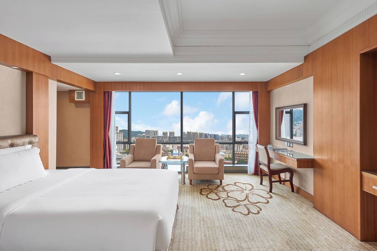 Habitación de hotel con una cama grande, dos sillones, un escritorio y un televisor. La habitación tiene ventanas amplias con vistas a la ciudad.