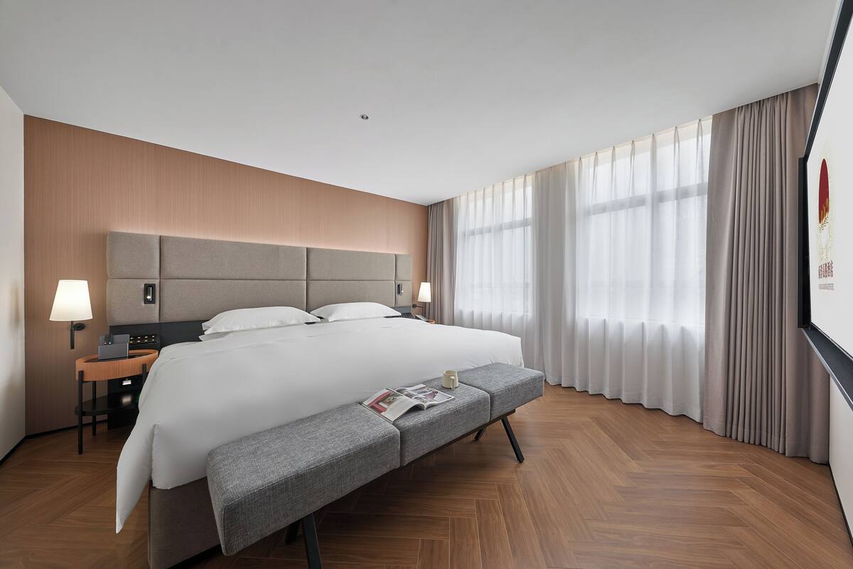 Una habitación de hotel moderna con una cama grande, mesitas de noche, un banco y vistas al exterior a través de amplios ventanales.