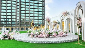 Un edificio moderno con una amplia fachada de vidrio y un espacio exterior circular con arreglos florales y una estructura blanca con arcos.
