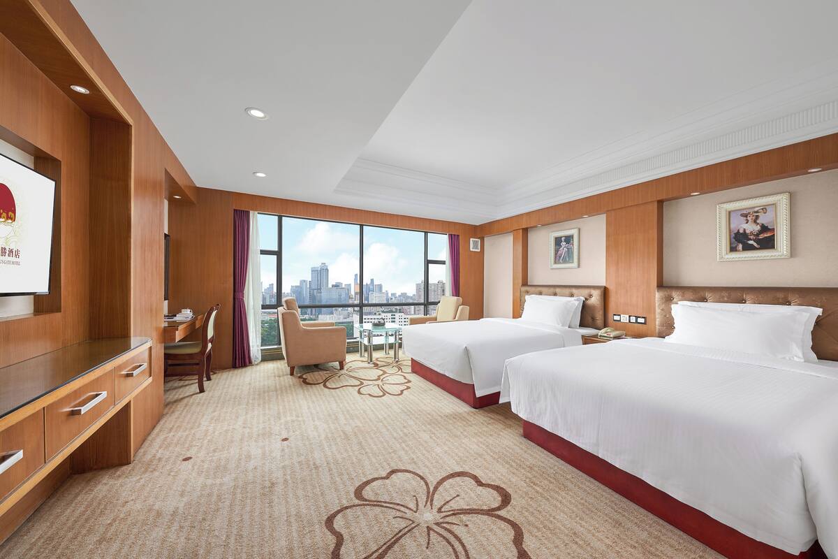 Habitación de hotel con dos camas, un escritorio, una silla y un amplio ventanal con vistas a la ciudad.