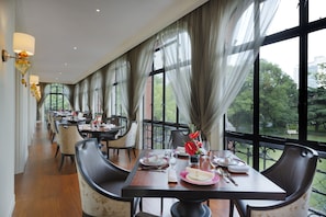 Un comedor con mesas dispuestas para una comida, ventanas amplias con cortinas y vistas al exterior con vegetación.