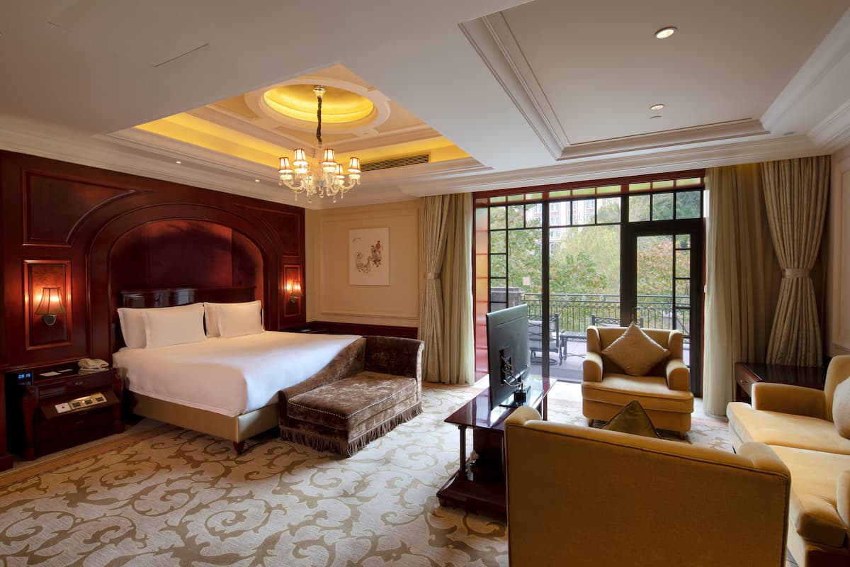 A hotel room with a large bed, a sitting area with a sofa and armchairs, a television, and a balcony with a view of greenery.