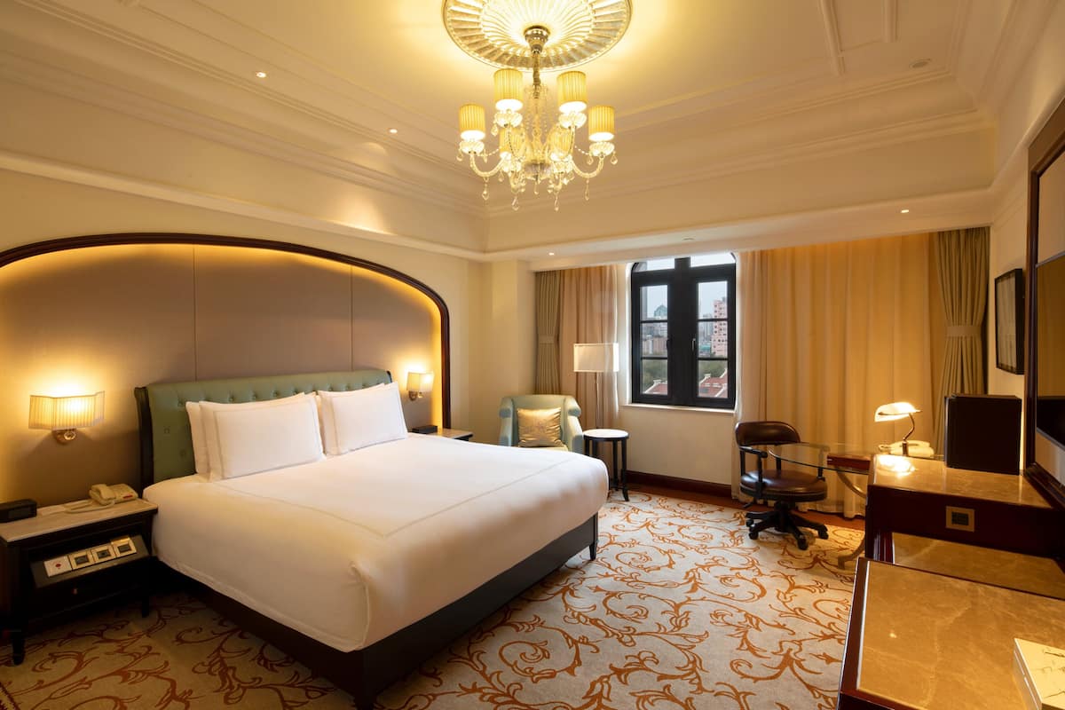 A hotel room with a large bed, a desk, a chair, a desk lamp, a telephone, and a window with curtains.