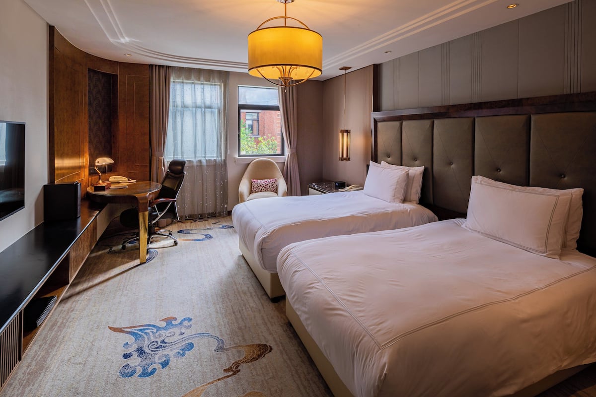 A hotel room with two beds, a desk with a chair, a television, and a large window.