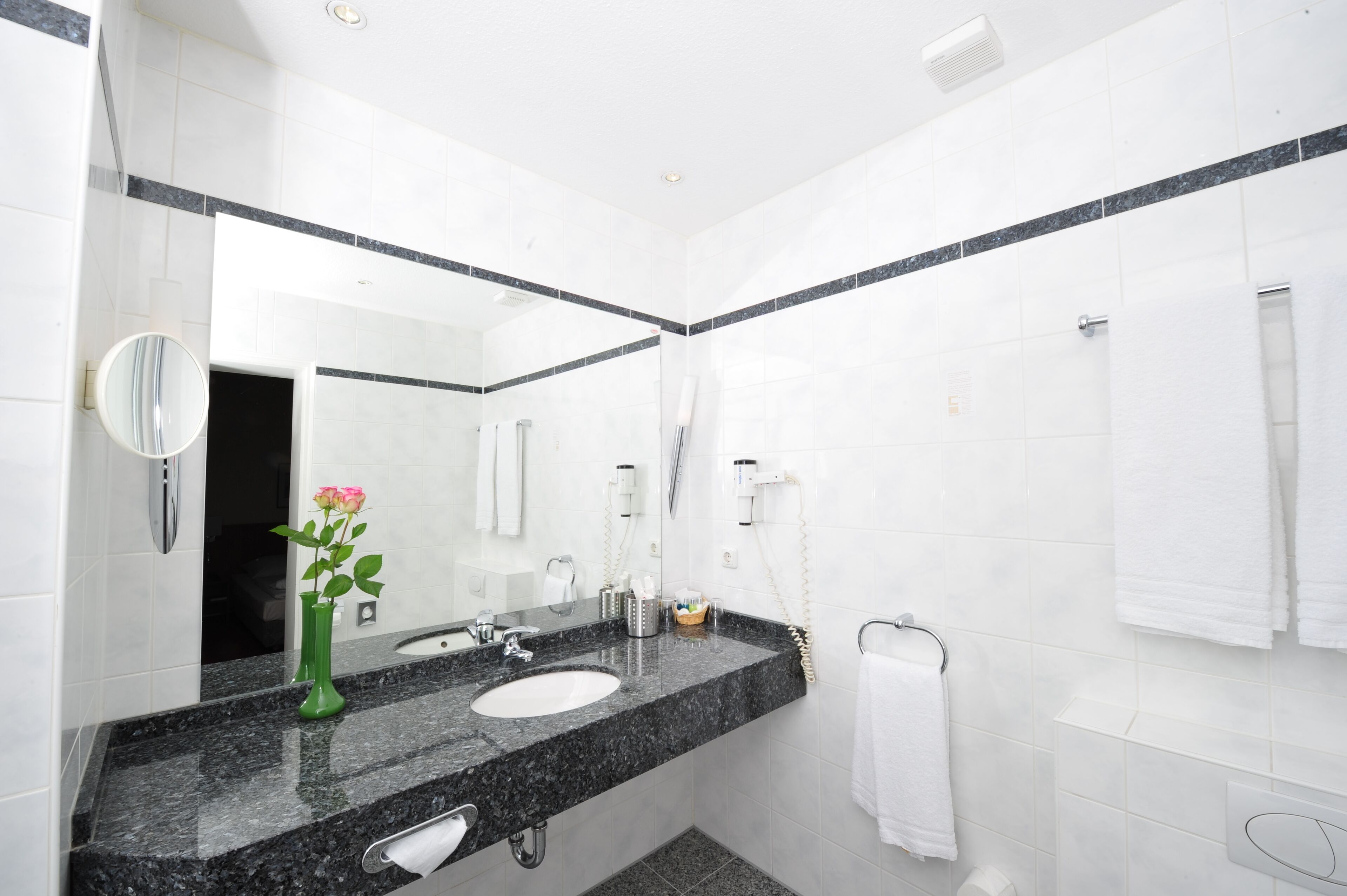 Ein modernes Badezimmer mit weißen Fliesen, einer Granitmarmorauskleidung, einem Waschbecken, einem Spiegel, einem Handtuchhalter und einer Vase mit einer Blume.