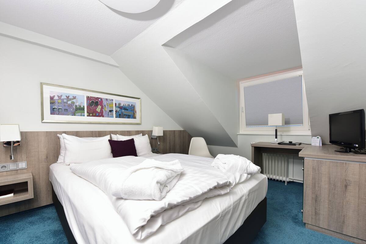 A hotel room with a bed, bedside tables, a desk with a computer, a TV, and a window with blinds.