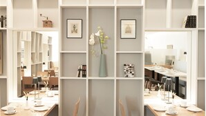 A dining area with two wooden tables set for two, each with chairs. Shelves with books and framed pictures are behind the tables. - Locals Stadthotel Bielefeld (Bielefeld)