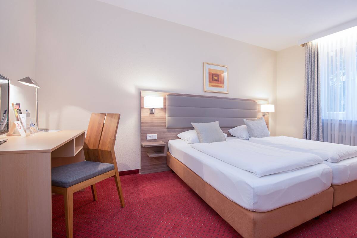 A hotel room with a bed, a desk, a chair, a nightstand, and a window with curtains.