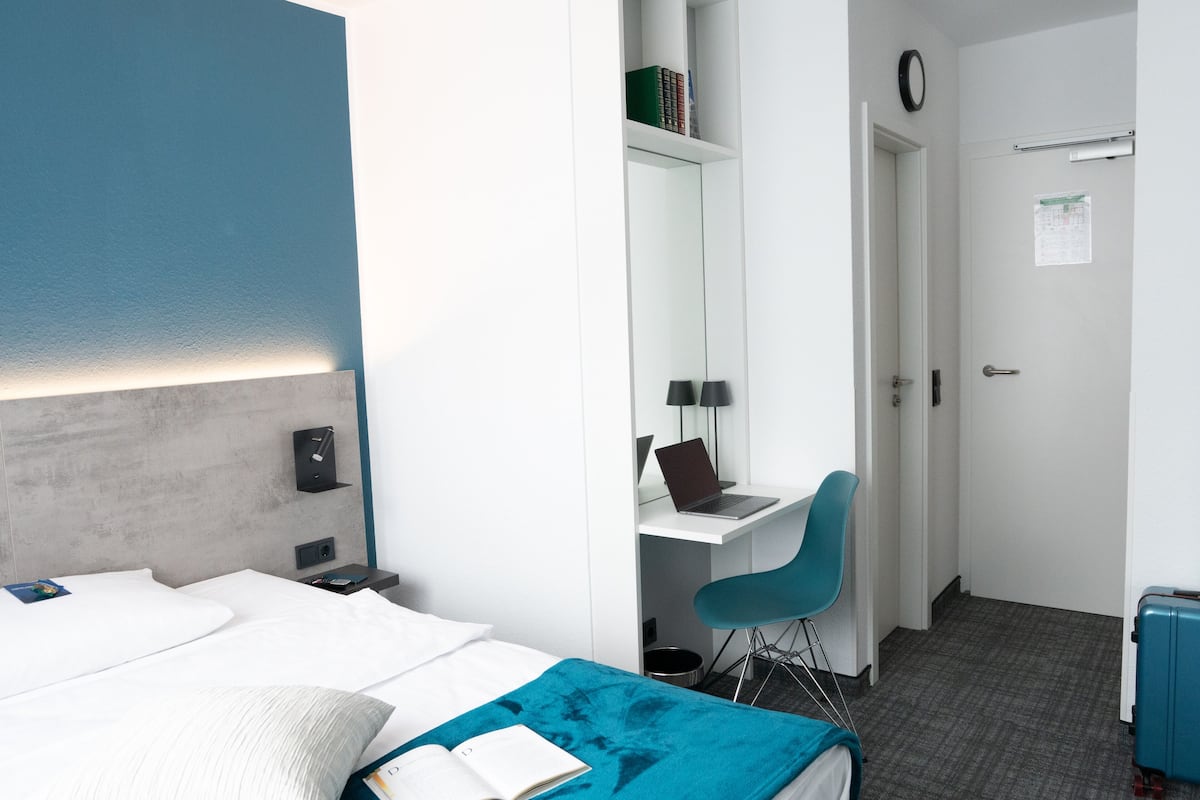 A modern hotel room with a bed, a desk, a chair, a laptop, and a suitcase.