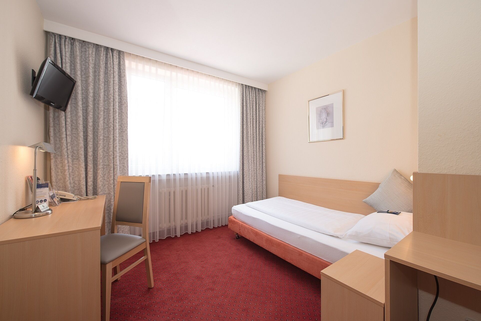 A hotel room with a bed, a desk with a chair, a television mounted on the wall, and a window with curtains.