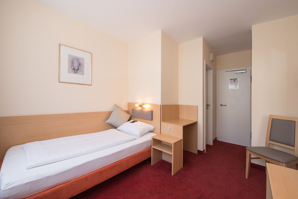 A hotel room with a single bed, a desk, a chair, a small table, a wall-mounted light, and a door.