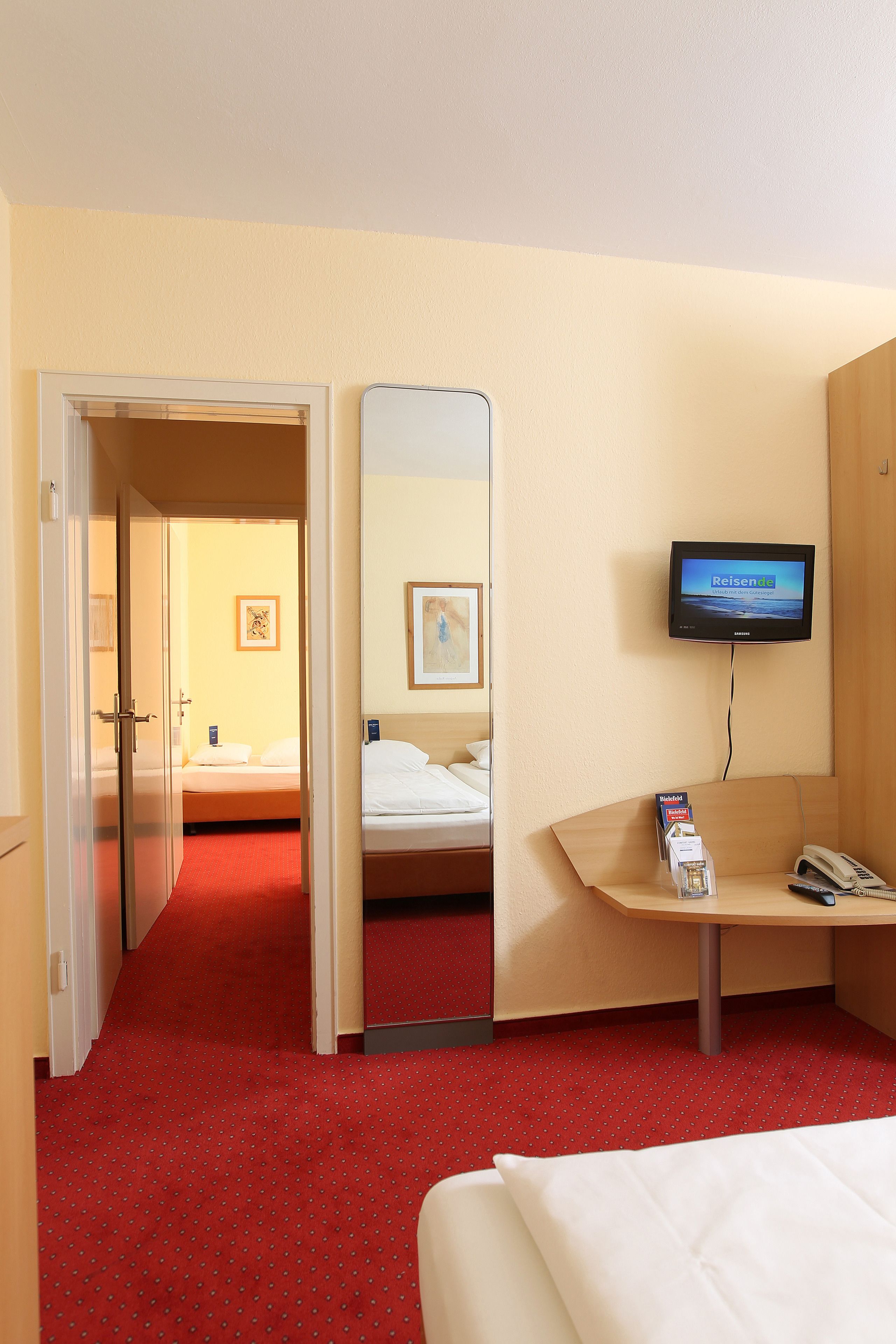 A hotel room with a bed, a desk with a phone and a magazine, a TV on the wall, and a mirror.