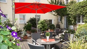 An outdoor patio with round tables and chairs, a red umbrella, and potted plants. - Locals Stadthotel Bielefeld (Bielefeld)