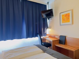 Habitación de hotel con cama, escritorio y silla, televisor montado en la pared y ventana con cortinas.