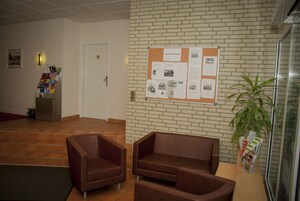 Eine Lobby mit einer Pinwand, zwei braunen Sofas, einem Topf mit Pflanze und einem Fliesenboden.