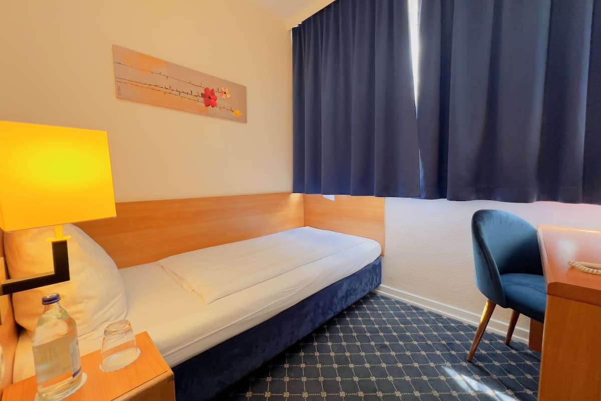 Habitación de hotel con una cama individual, una mesita de noche con lámpara, una silla y un ventanal con cortinas.