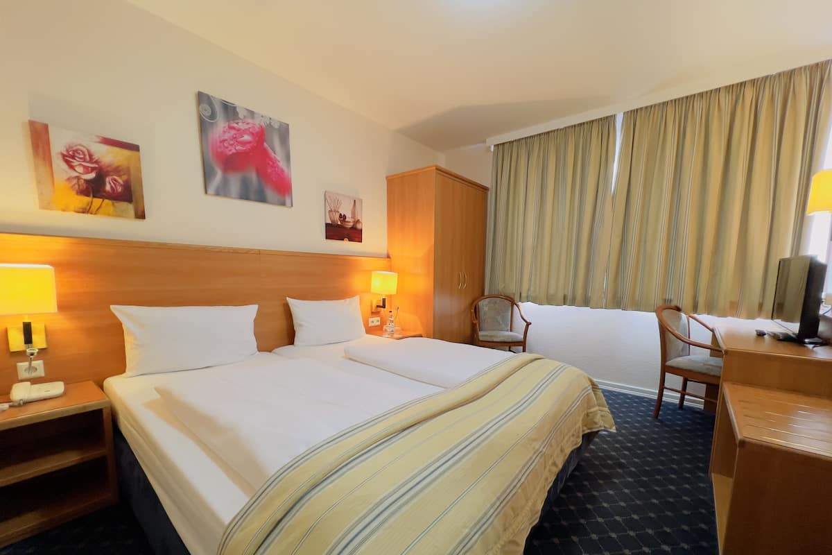 Habitación de hotel con una cama grande, un escritorio con computadora, una silla y un televisor. Hay dos lámparas en las mesitas de noche y una ventana con cortinas.