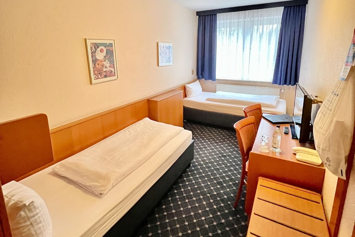 Habitación de hotel con dos camas individuales, un escritorio con silla, una mesita y una ventana con cortinas.