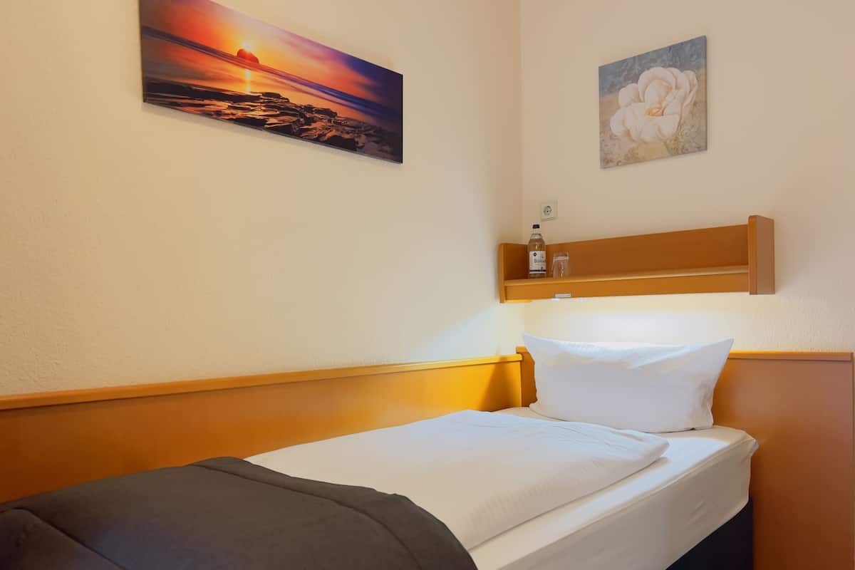 Una cama bien hecha con cabecera, un estante montado en la pared con una botella y un vaso, y dos cuadros.