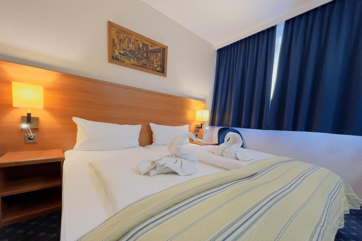 Habitación de hotel con una cama grande, dos almohadas, cabecera de madera, un cuadro en la pared, una mesita de noche, una lámpara y una ventana con cortinas.
