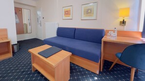 A hotel room with a blue sofa, a wooden desk, a small table, a chair, a lamp, and two framed pictures on the wall.