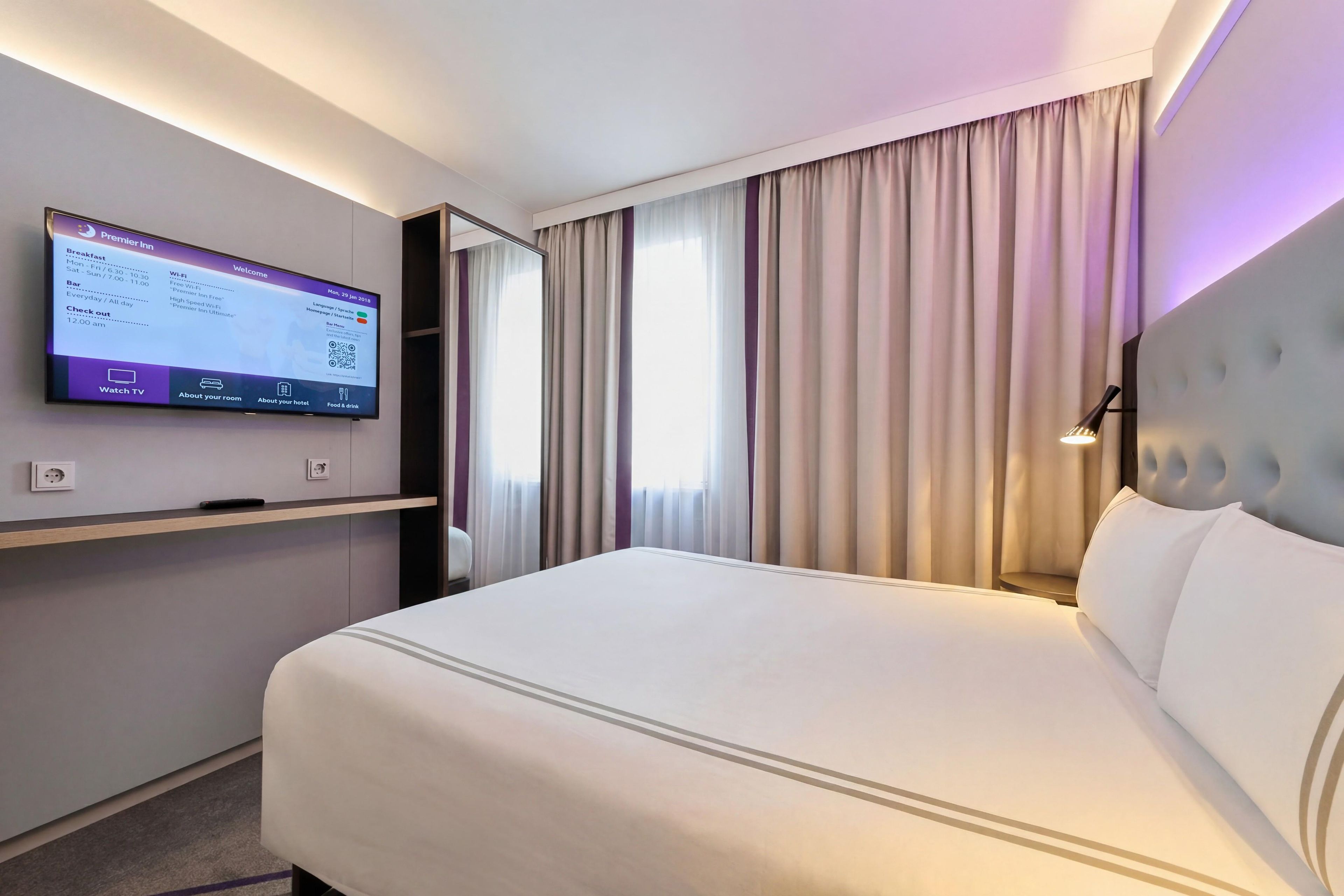 A hotel room with a bed, a TV mounted on the wall, and a window with curtains.