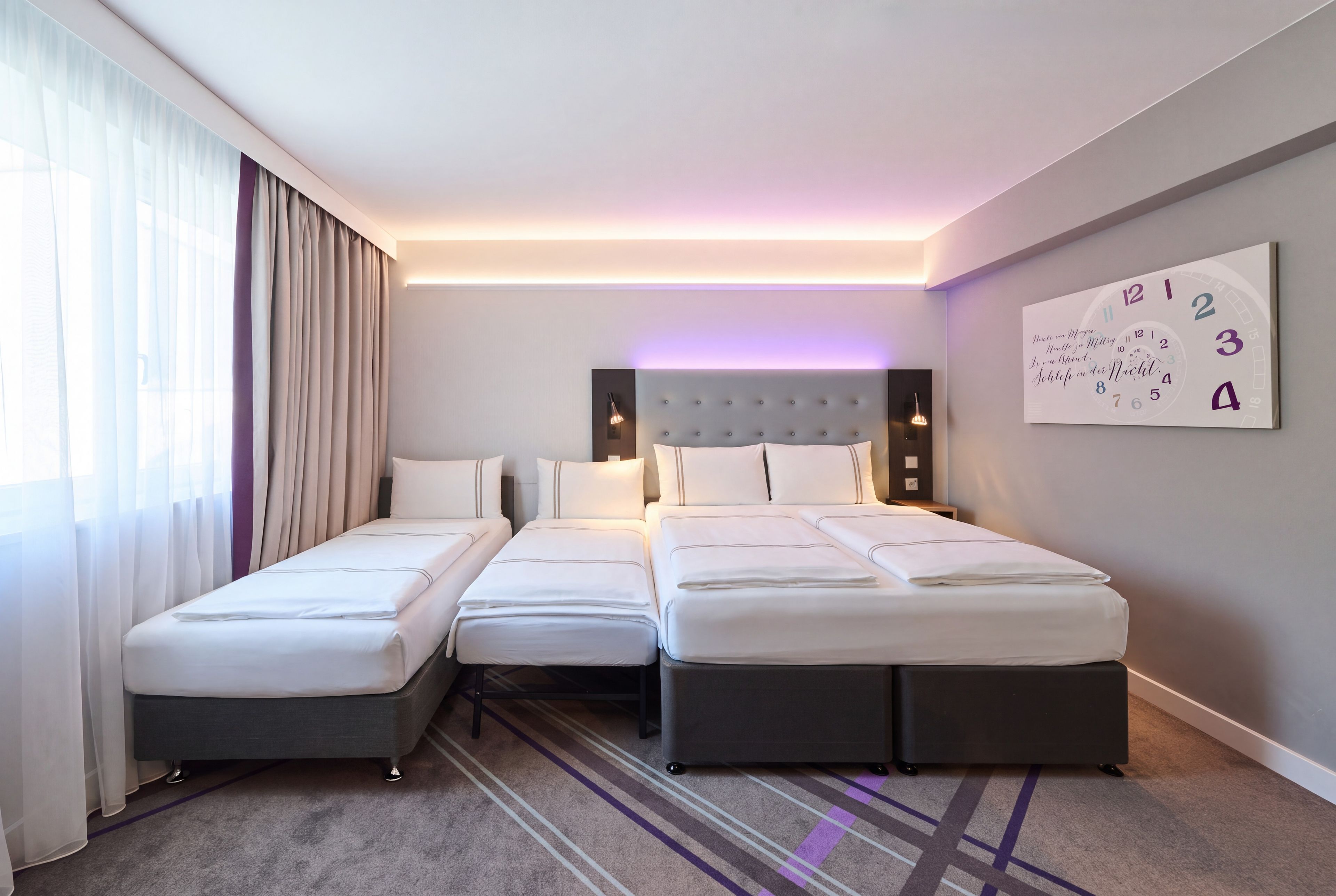A hotel room with two single beds and one double bed, a large window with curtains, a wall clock, and a carpeted floor.