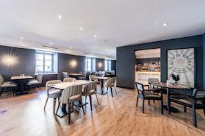 Et moderne spiseområde med flere borde og stole, et køkken i baggrunden, samt en væg med solstråle-design.