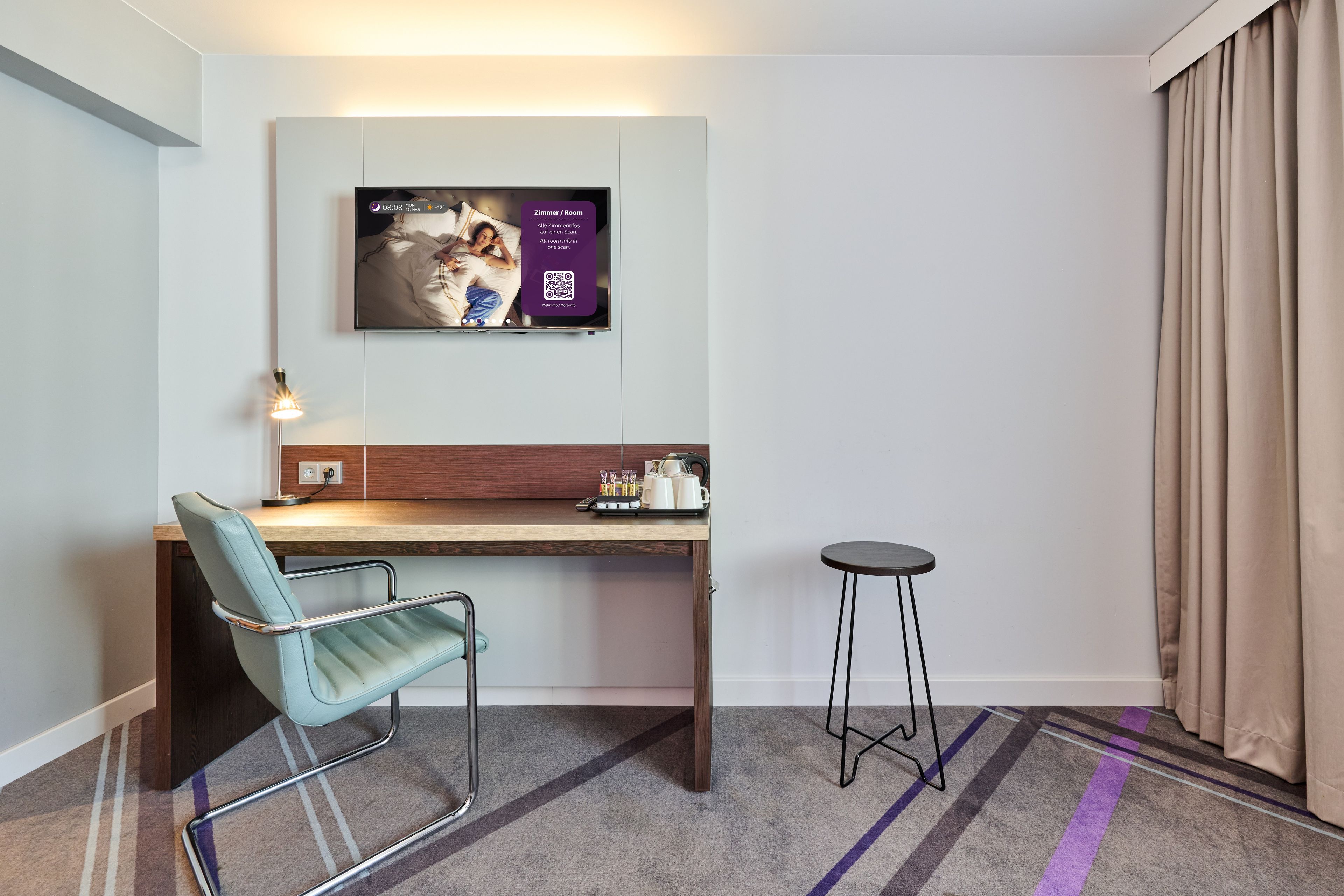 A hotel room with a bed, a desk, a chair, a small stool, and a television mounted on the wall.