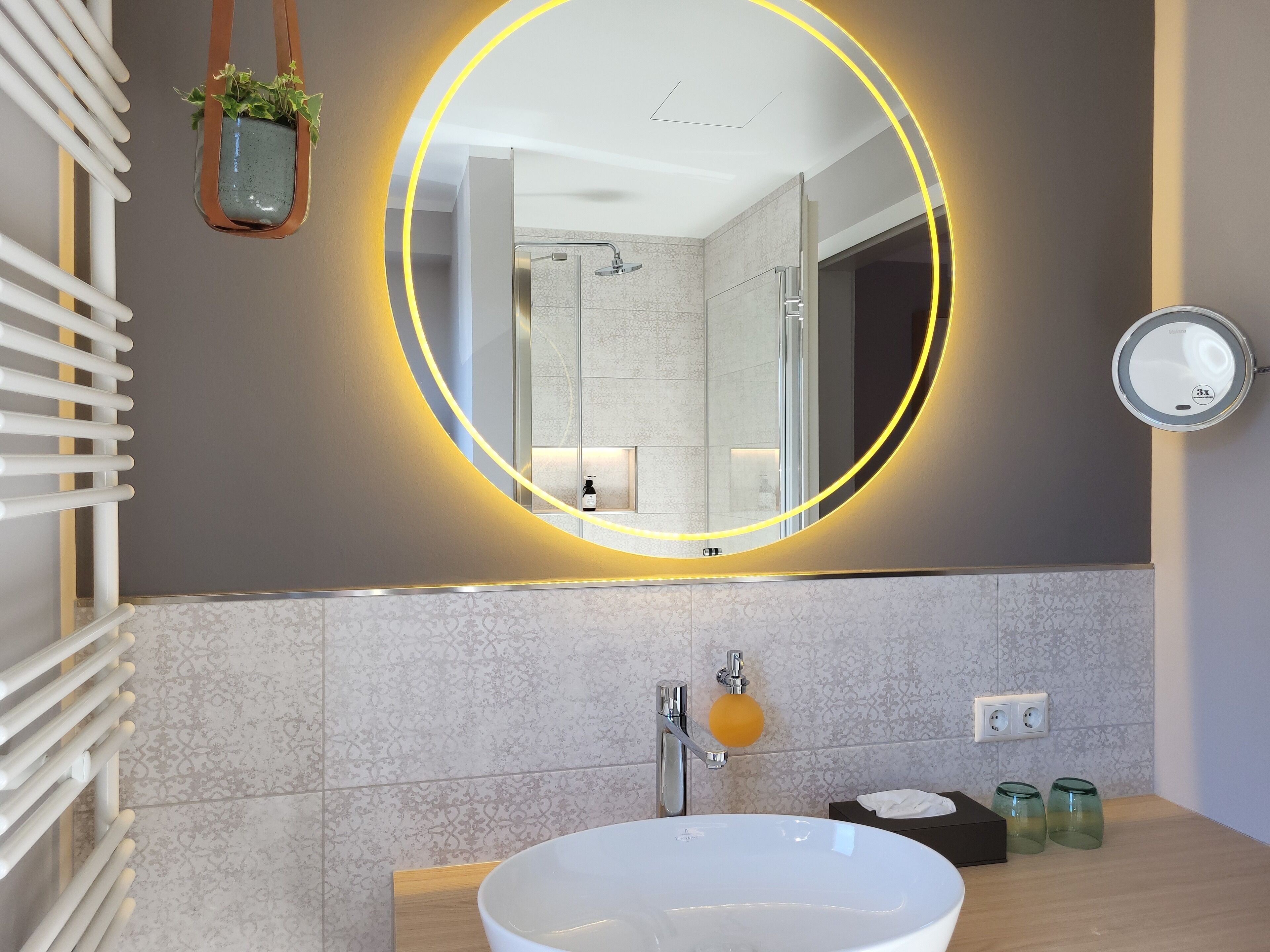 A modern bathroom with a large oval mirror, a white sink, and a glass shower enclosure.