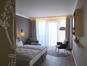 A modern hotel room with a large bed, two chairs, a round rug, and a large window with curtains.
