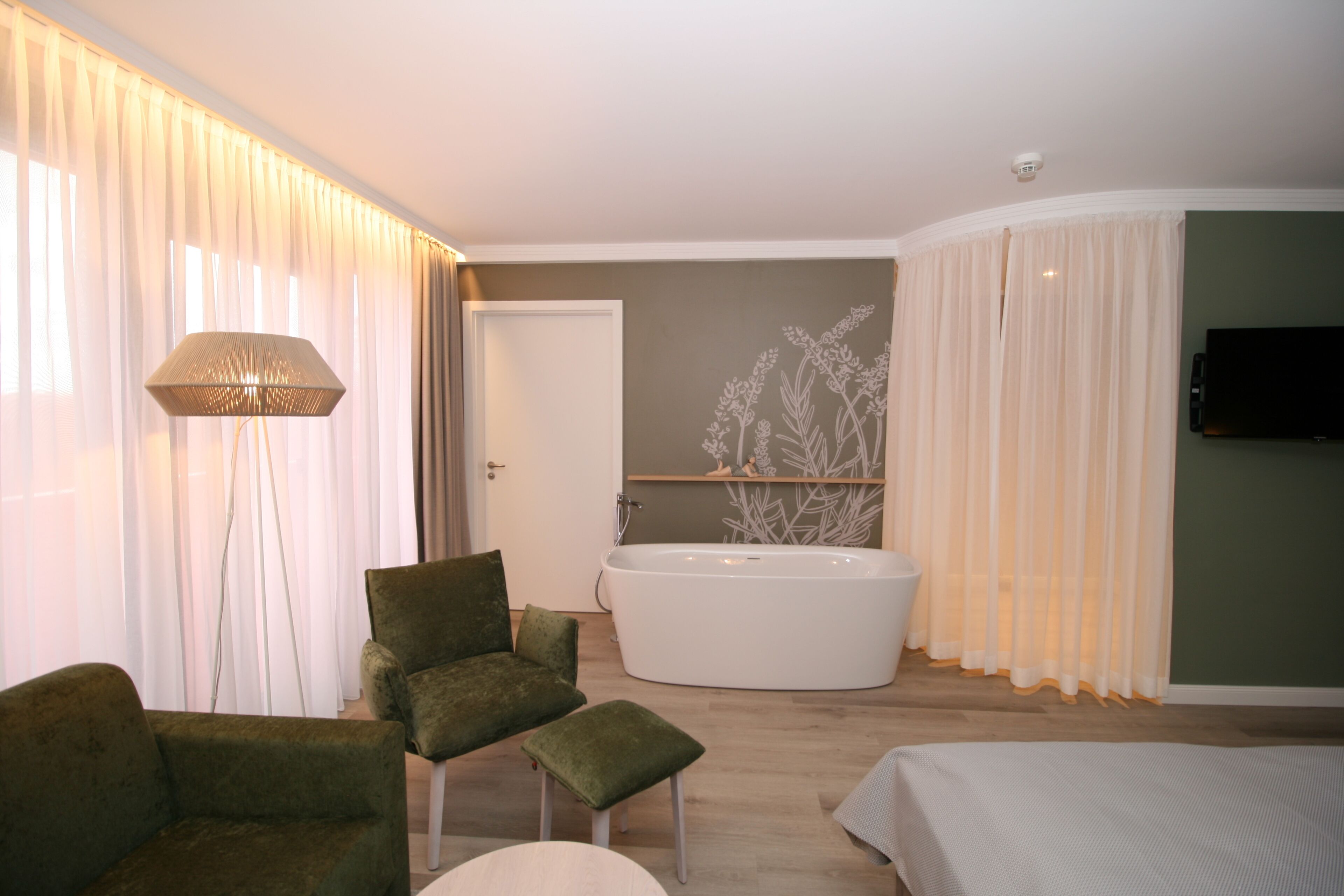 Een moderne slaapkamer met een groot bed, een groene fauteuil, een klein krukje, een vloerlamp, een groot raam met dunne gordijnen, een badkuip en een flatscreen televisie aan de muur.