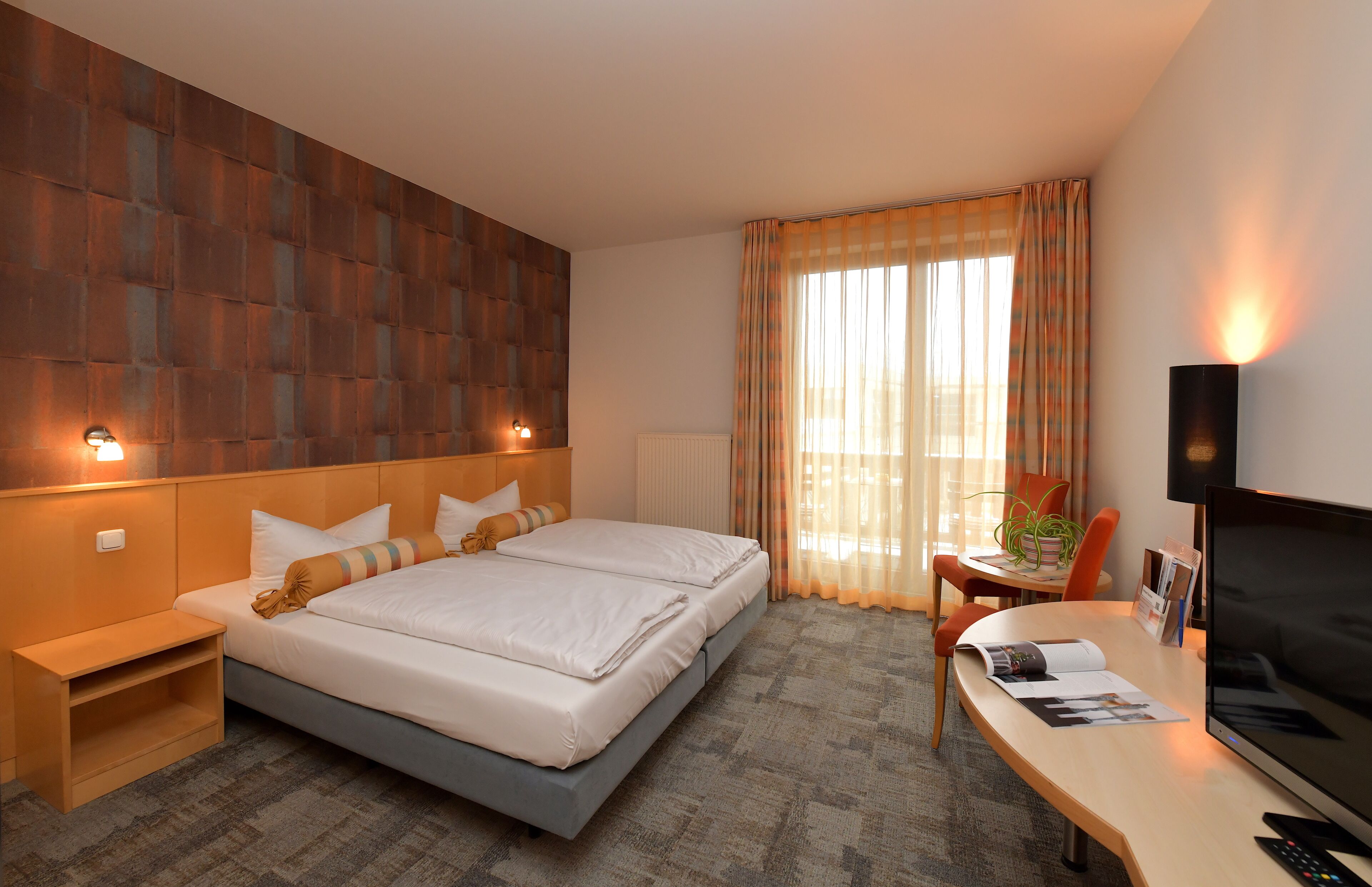 A hotel room with a large bed, a desk with a TV, a chair, and a window with curtains.