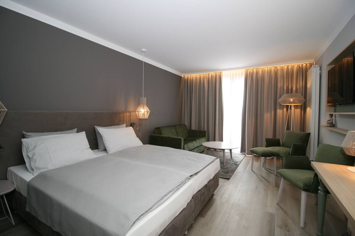 A modern hotel room with a large bed, a small round table, green armchairs, and a sofa.