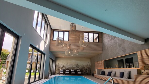 A modern indoor swimming pool with a clear view of the sky through large windows, a poolside seating area, and a cityscape mural on the wall. - Hotel Rittmeister (Rostock)