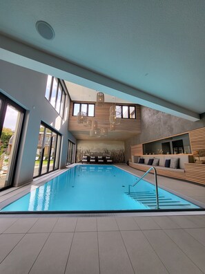 Une piscine intérieure moderne, dotée de grandes baies vitrées offrant une vue dégagée sur le ciel, d’un espace de détente au bord de la piscine et d’une fresque représentant un paysage urbain sur le mur.