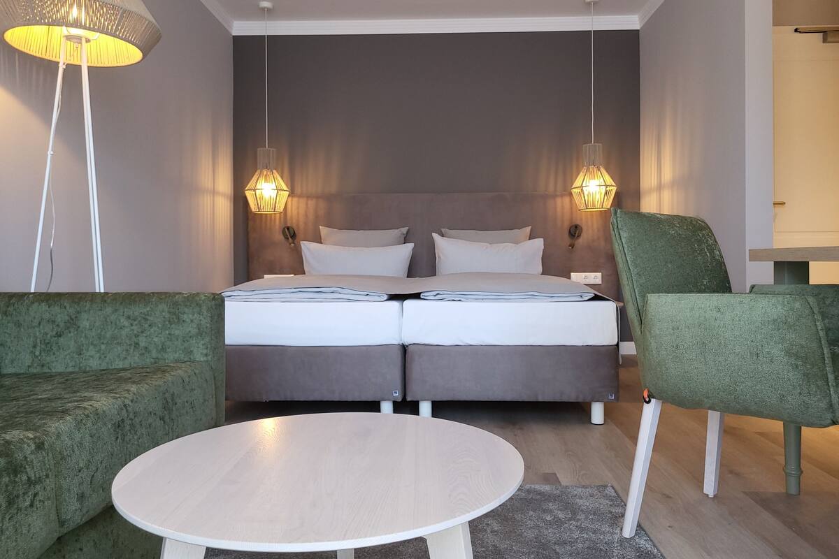 A modern hotel room with a double bed, two bedside lamps, a green armchair, a round white table, and a floor lamp.