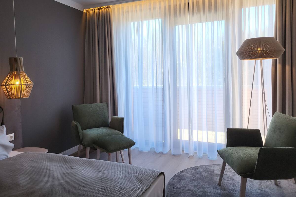 A hotel room with a bed, two armchairs, a standing lamp, and a large window with sheer curtains.
